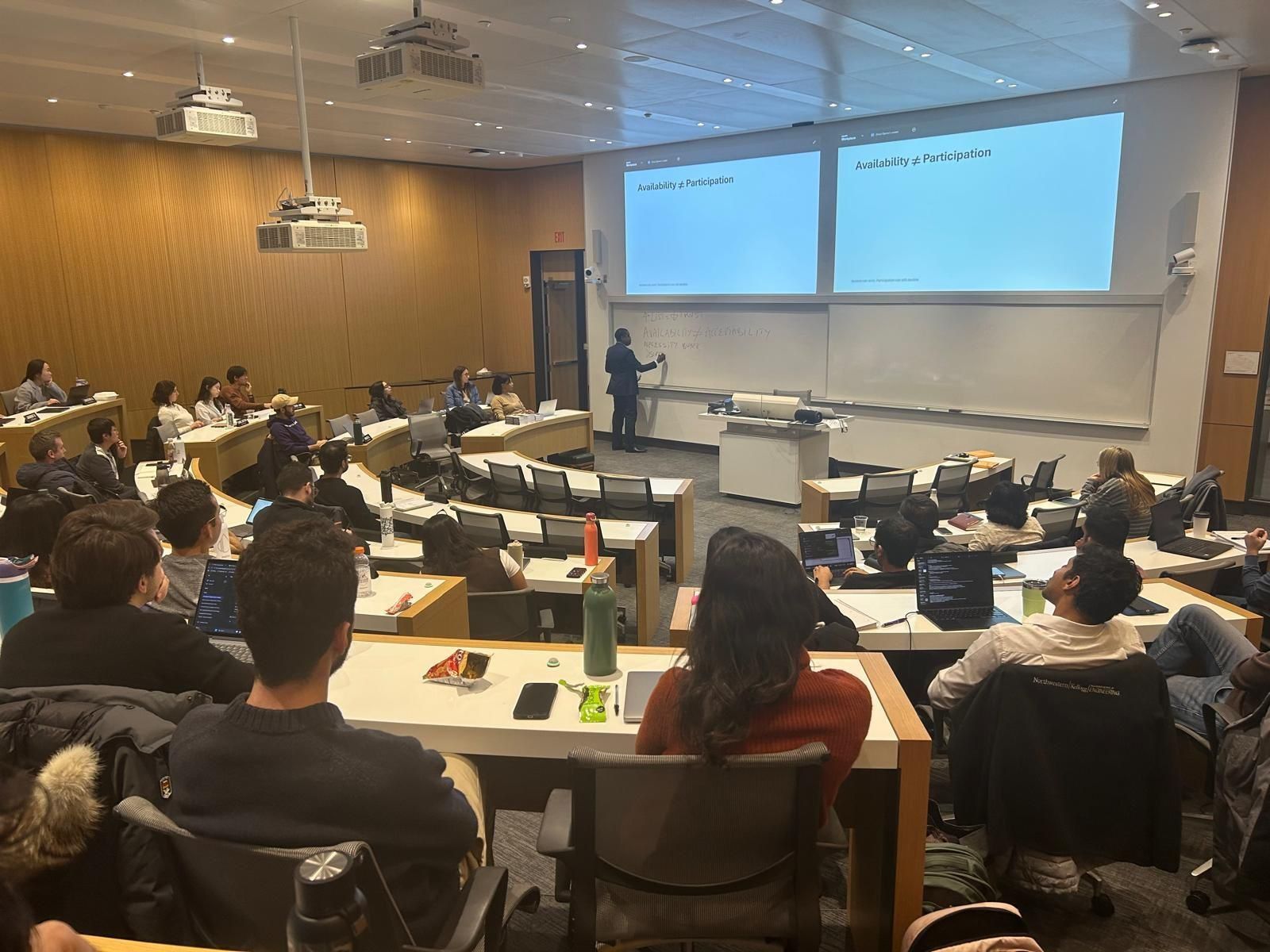 Akin presenting at the front of a lecture theatre, students at their desks with laptops.